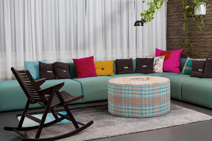 A modern living space features a curved turquoise sofa adorned with colorful cushions, a round checkered coffee table, a rocking chair, and a potted plant hanging from the ceiling, all set against sheer white curtains.